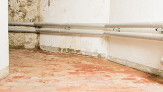 Verfärbte Kellerwand mit Leitungsrohren über Ziegelboden.